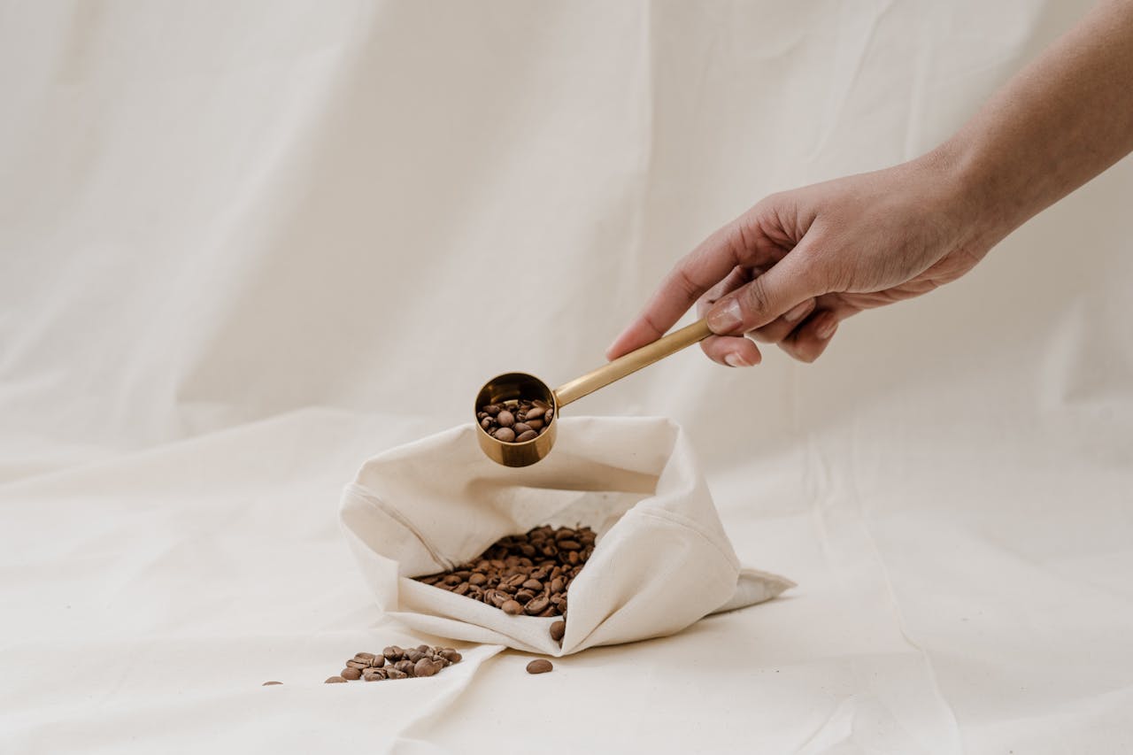 A hand holding a scoop over a fabric bag filled with coffee beans, highlighting a minimalist aesthetic.